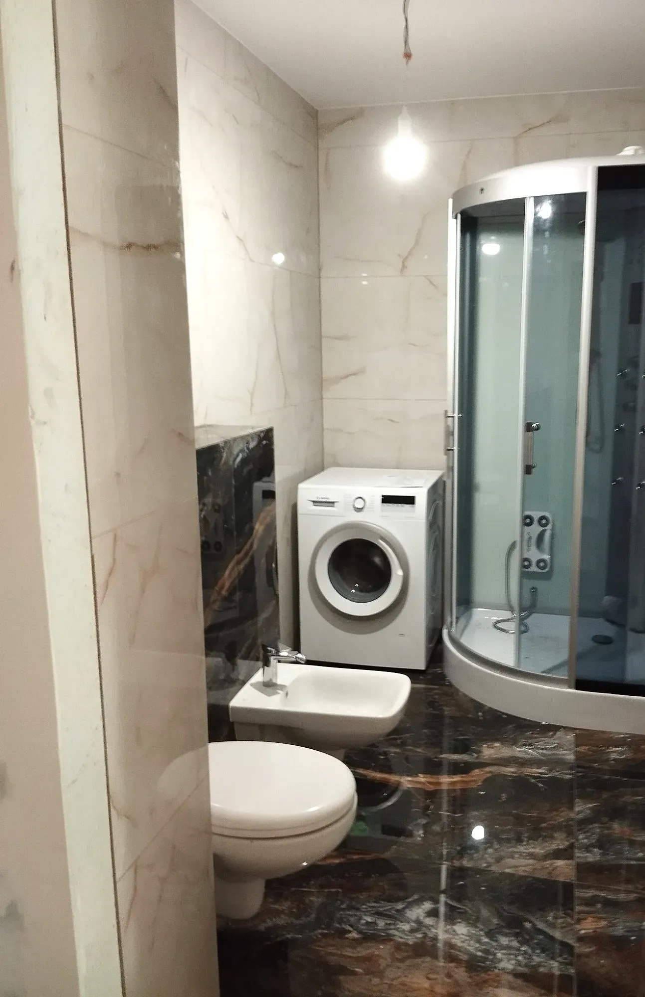 Ta sama łazienka po remoncie: jasne płytki imitujące marmur, ciemna podłoga, kabina prysznicowa, podwieszana toaleta i bidet